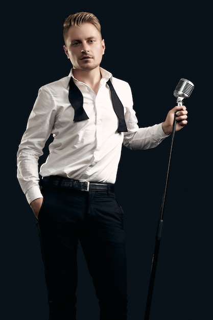 Portrait of handsome blond man singer in elegant tuxedo and bow tie posing with vintage microphone on black  wall