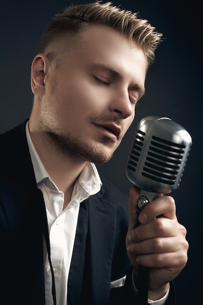 Portrait of handsome blond man singer in elegant tuxedo and bow tie posing with vintage microphone on black  wall