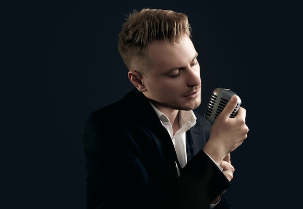 Portrait of handsome blond man singer in elegant tuxedo and bow tie posing with vintage microphone on black  wall