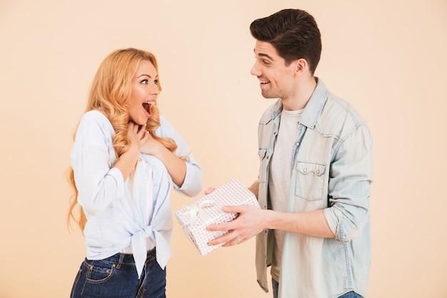 Portrait of handsome brunette man giving present box to excited happy woman, isolated over beige wall