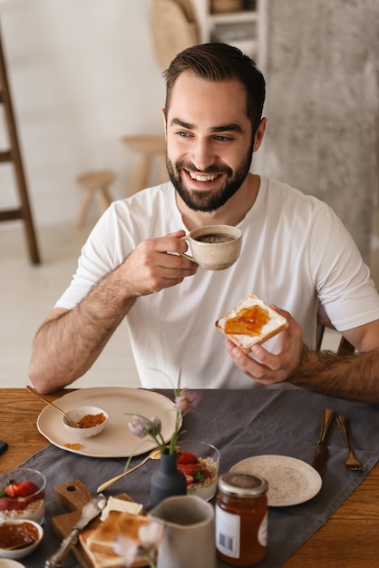 Portrait of handsome brunette man  using smartphone while having breakfast in stylish kitchen at home