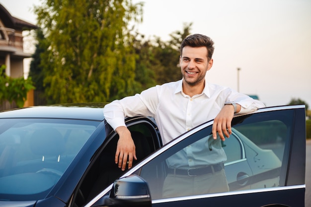 Portrait of handsome businesslike man wearing suit, standing near his luxury black car with open driver's door