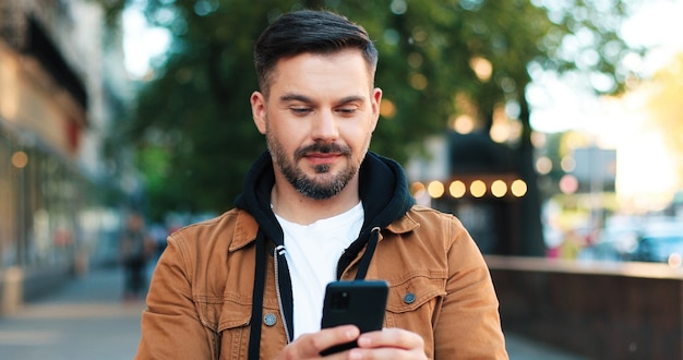 Portrait of the handsome caucasian man showing charming smile and healthy teeth while walking with his smartphone at the street. People and gadgets concept