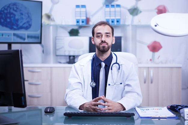 Portrait of handsome and confident doctor looking confident in the camera in his cabinet. Young surgeon with white coat.