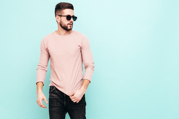 Portrait of handsome confident model Sexy stylish man dressed in pink sweater and jeans Fashion hipster male posing near blue wall in studio In sunglasses Isolated