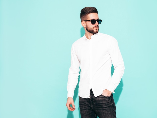 Portrait of handsome confident model Sexy stylish man dressed in white shirt and jeans Fashion hipster male posing near blue wall in studio Isolated In sunglasses