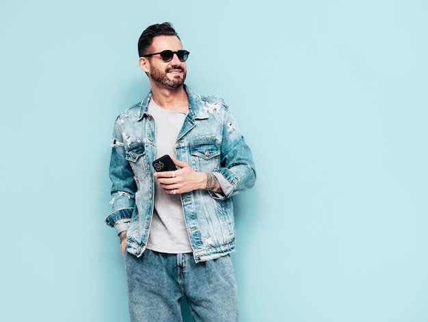 Portrait of handsome confident stylish hipster lambersexual model Sexy man dressed in jeans jacket Fashion male isolated on blue wall in studio In sunglasses