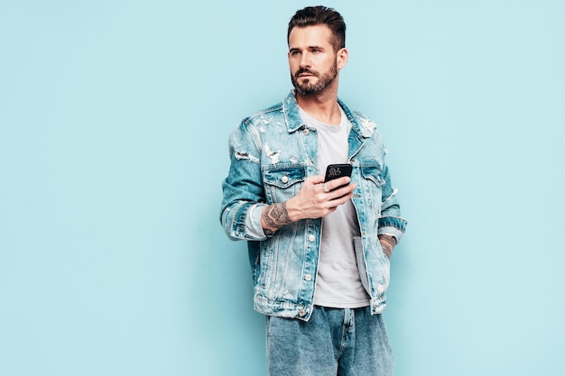 Portrait of handsome confident stylish hipster lambersexual model Sexy man dressed in jeans jacket Fashion male isolated on blue wall in studio