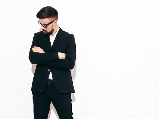 Portrait of handsome confident stylish hipster lambersexual model Sexy modern man dressed in elegant black suit Fashion male posing in studio near white wall Isolated on white In spectacles