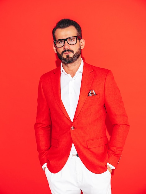 Portrait of handsome confident stylish hipster lambersexual model Sexy modern man dressed in elegant suit Fashion male posing in studio near red wall in spectacles