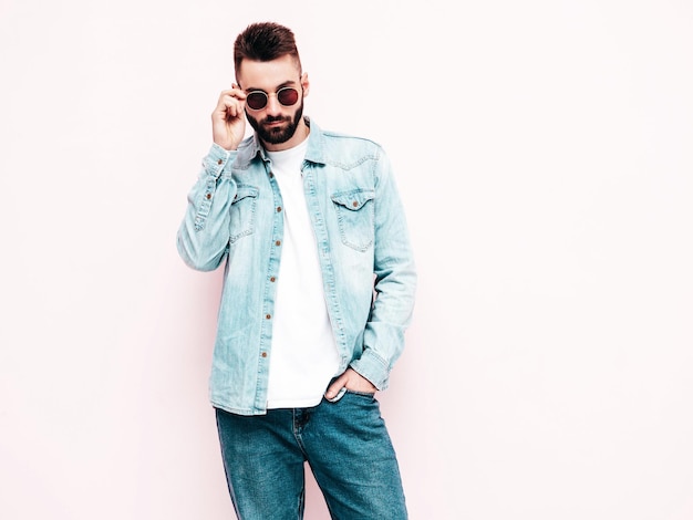 Portrait of handsome confident stylish hipster lambersexual modelMan dressed in jacket and jeans Fashion male posing in studio in sunglasses Isolated on light pink