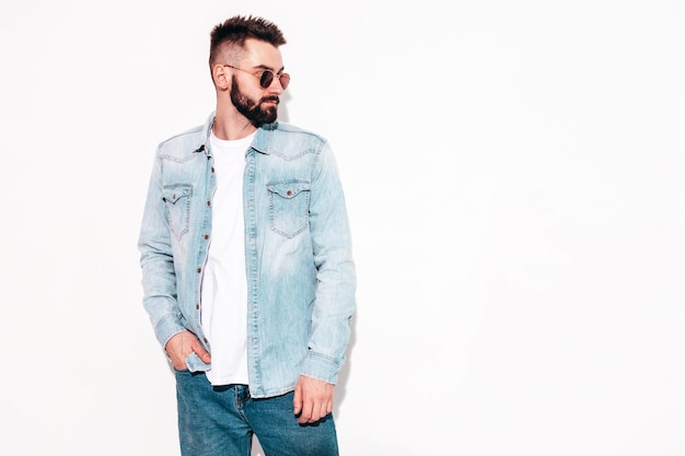 Portrait of handsome confident stylish hipster lambersexual modelMan dressed in jacket and jeans Fashion male posing in studio in sunglasses Isolated on white