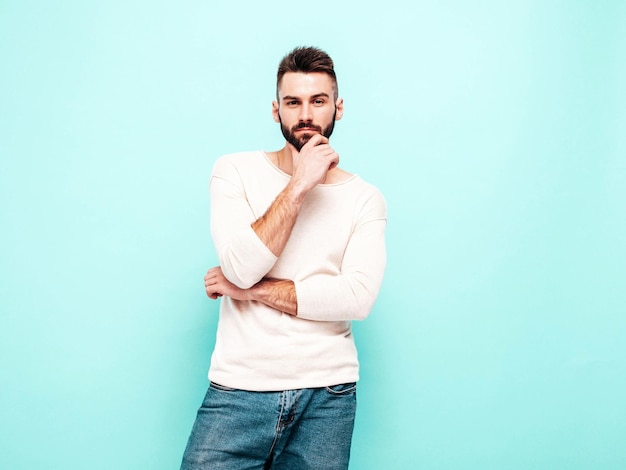 Portrait of handsome confident stylish hipster lambersexual modelMan dressed in white sweater and jeans Fashion male isolated on blue wall in studio Thoughtful