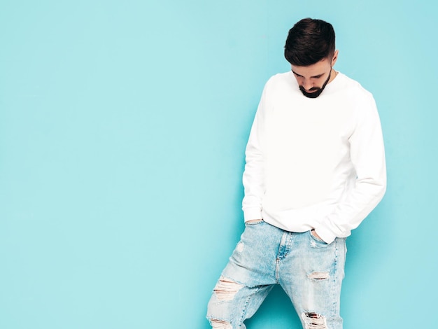 Portrait of handsome confident stylish hipster lambersexual modelMan dressed in white sweater and jeans Fashion male isolated on blue wall in studio