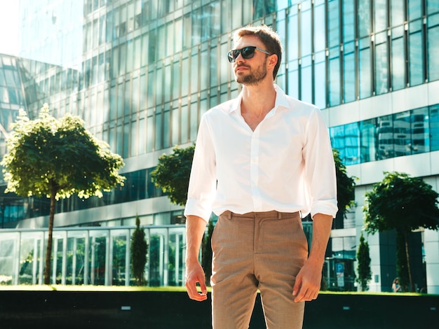 Portrait of handsome confident stylish hipster lambersexual modelModern man dressed in white shirt Fashion male posing in the street background near skyscrapers in sunglasses