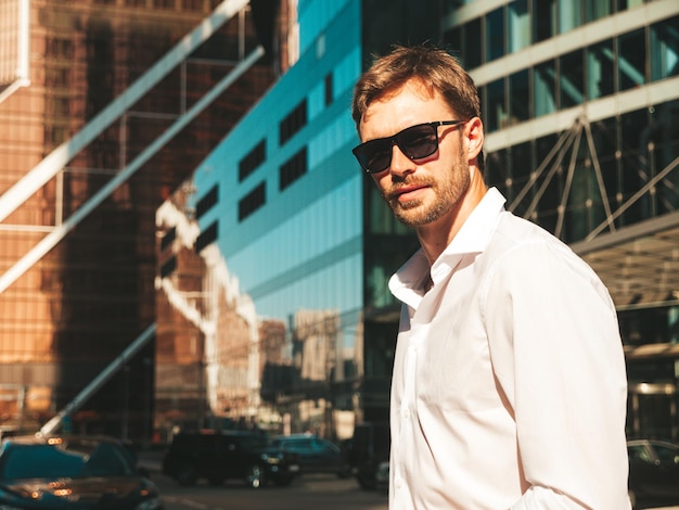 Portrait of handsome confident stylish hipster lambersexual modelModern man dressed in white shirt Fashion male posing in the street background near skyscrapers in sunglasses