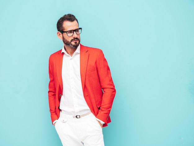 Portrait of handsome confident stylish hipster lambersexual modelSexy modern man dressed in elegant red suit Fashion male posing in studio near blue wall in spectacles