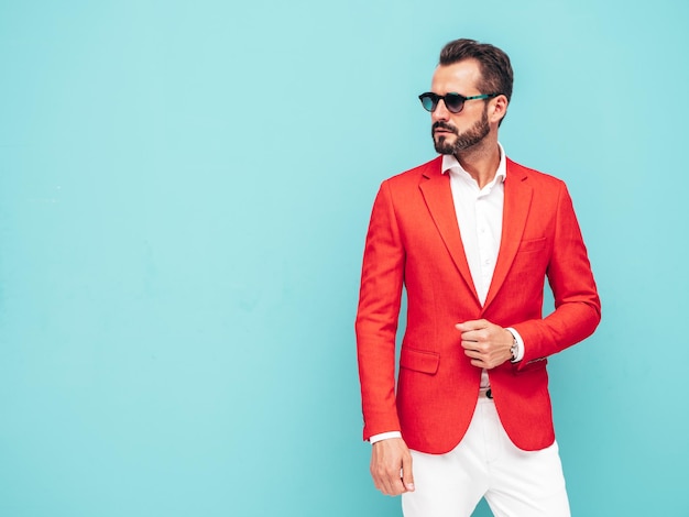 Portrait of handsome confident stylish hipster lambersexual modelSexy modern man dressed in elegant red suit Fashion male posing in studio near blue wall in sunglasses