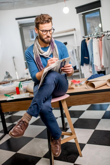 Portrait of a handsome fashion designer sitting with clothing sketches at the studio full of tailoring tools and clothes