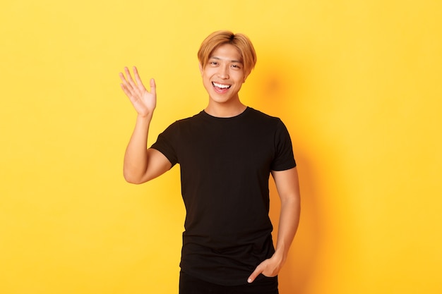 Portrait of handsome friendly asian guy in black outfit, waving hand to say hello and smiling, greeting someone, standing yellow wall