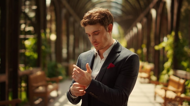 Portrait of handsome groom looking at camera under arch in park Smiling man buttoning up shirt in park Attractive guy getting ready for wedding ceremony outdoors