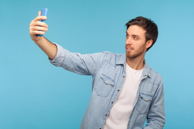 Portrait of handsome happy man with stylish haircut in denim shirt talking on video call, having online conversation on mobile phone, taking selfie. indoor studio shot isolated on blue background