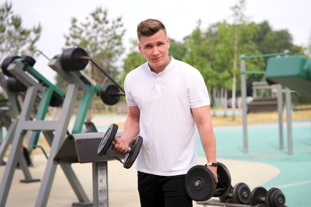 Portrait of handsome happy strong athletic man athlete bodybuilder training with dumbbells outdoors at open air gym at summer gain hands and shoulders muscles Bodybuilding fitness healthy lifestyle