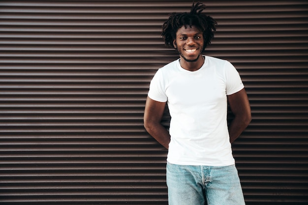 Portrait of handsome hipster modelUnshaven African man dressed in white summer tshirt and jeans Fashion male with dreadlocks hairstyle posing near roller shutter wall in the street