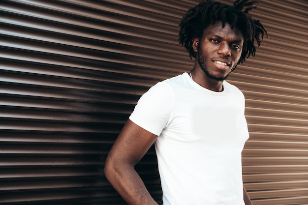 Portrait of handsome hipster modelUnshaven African man dressed in white summer tshirt and jeans Fashion male with dreadlocks hairstyle posing near roller shutter wall in the street