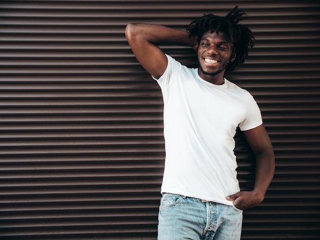 Portrait of handsome hipster modelUnshaven African man dressed in white summer tshirt and jeans Fashion male with dreadlocks hairstyle posing near roller shutter wall in the street