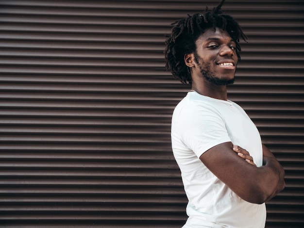 Portrait of handsome hipster modelUnshaven African man dressed in white summer tshirt and jeans Fashion male with dreadlocks hairstyle posing near roller shutter wall in the street