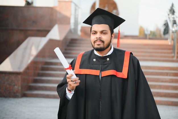 Portrait of handsome indian graduate student in graduation glow with diploma