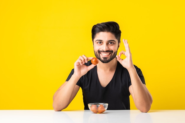 Portrait of a handsome Indian young manÃÂ eating sweet Gulab Jamun against yellow background