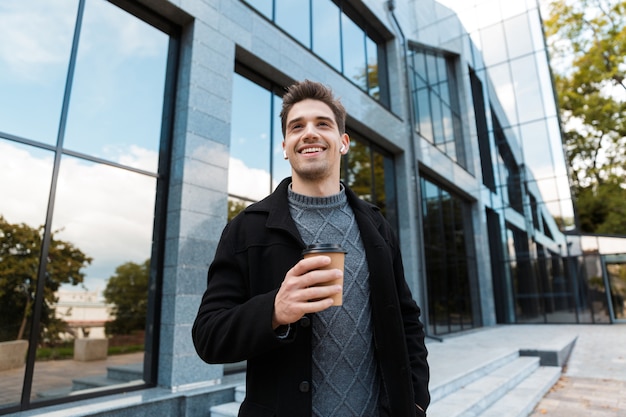 Portrait of handsome man 30s wearing earpods holding paper cup of takeaway coffee while strolling in front of glass building