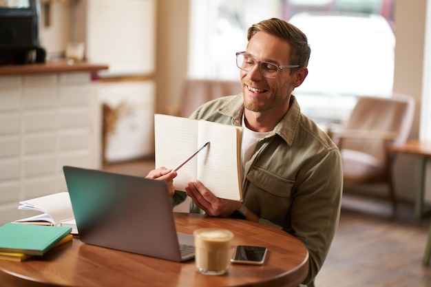 Portrait of handsome man in glasses working remotely from empty cafe showing example in his notebook