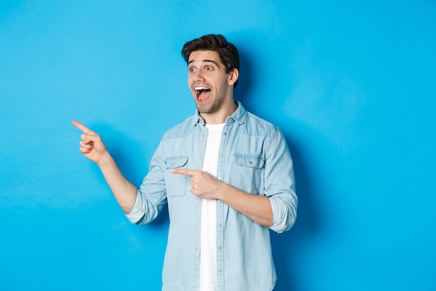 Portrait of handsome man looking and pointing fingers left with excitement, standing against blue wall