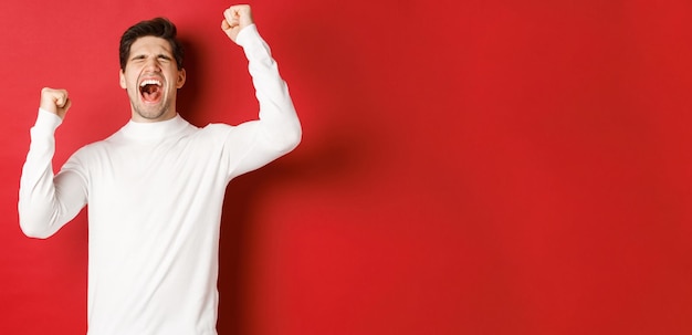 Portrait of handsome man in white sweater feeling cheerful celebrating victory shouting for joy and