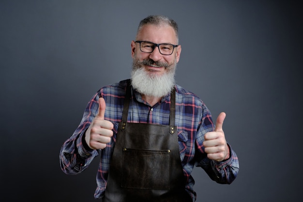 Portrait of handsome mature bearded man dressed leather apron gesturing thumbs up over gray wall, caucasian workman with beard smiling