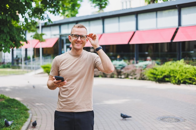 Portrait of handsome mature businessman holding messenger bag wearing stylish eyeglasses looking at camera Smiling middle aged man using smartphone standing on the street Mobile banking concept