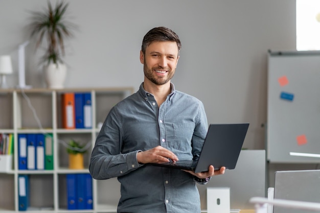 Portrait of handsome mature male office worker with laptop in hands standing in office and smiling