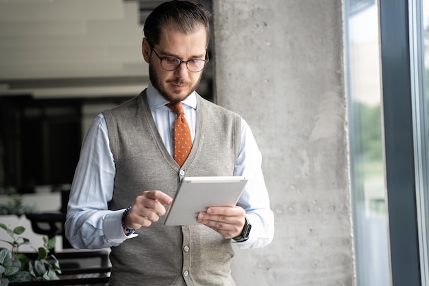 Portrait of handsome middleaged man holding digital tablet and smiling happily at camera standing by window in modern office