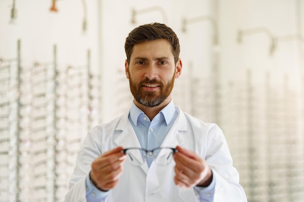 Portrait of handsome ophthalmologist offering glasses to client in optics store