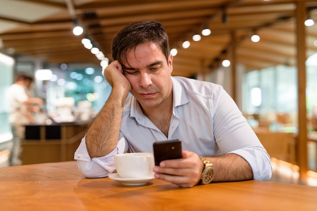 Portrait of handsome Persian man sitting at coffee shop while using mobile phone