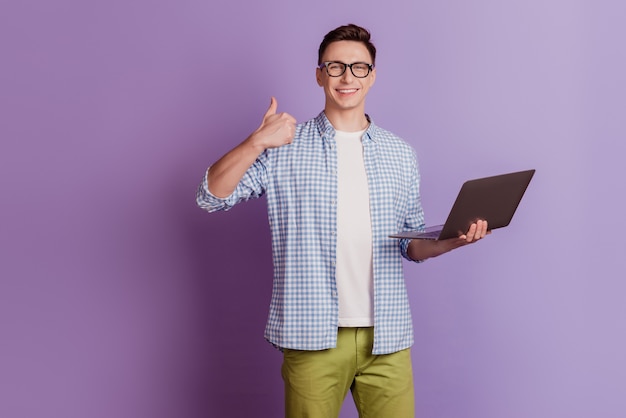 Portrait of handsome programmer guy hold netbook raise thumb up on violet background