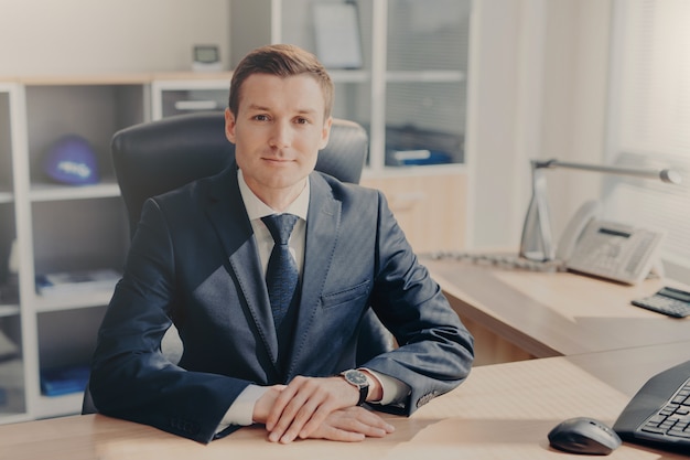 Portrait of handsome prosperous male manager sits at cabinet, looks directly into camera