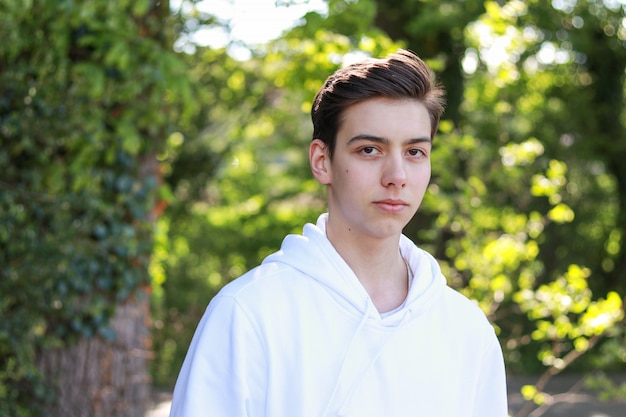 portrait of handsome serious teenager boy in white hoody in park outdoors at bright sunny day.