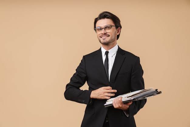 Portrait of a handsome smiling confident young businessman wearing suit standing isolated over beige wall, holding stack of documents