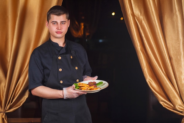 Portrait of handsome smiling male chef cook in uniform standing with delicious dish at the kitchen. Male chef holding plate with cooked dish.