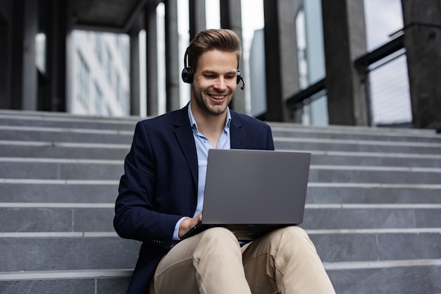 Portrait of handsome smiling man in casual wear sitting on bench using laptop for online meeting vid