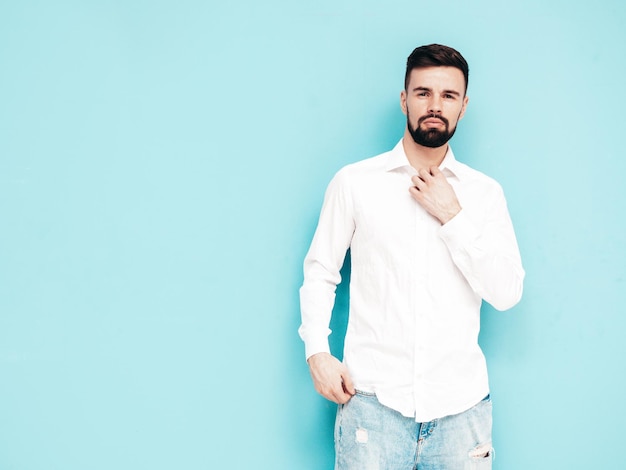 Portrait of handsome smiling model Sexy stylish man dressed in white shirt and jeans Fashion hipster male posing near blue wall in studio Isolated